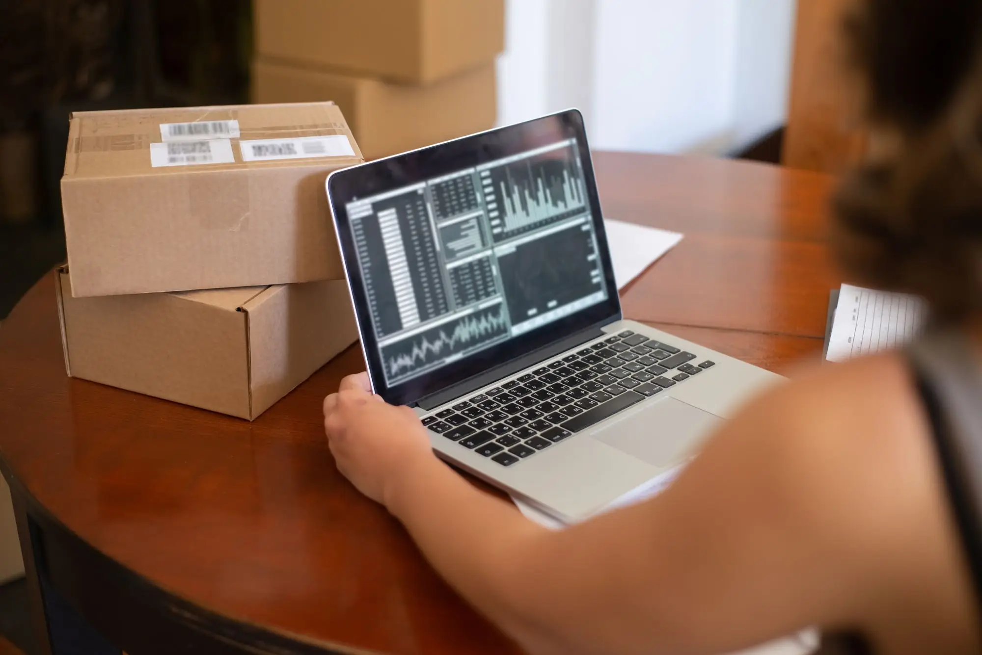 Online store owner checking sales and payout charts on a laptop beside labelled shipping boxes